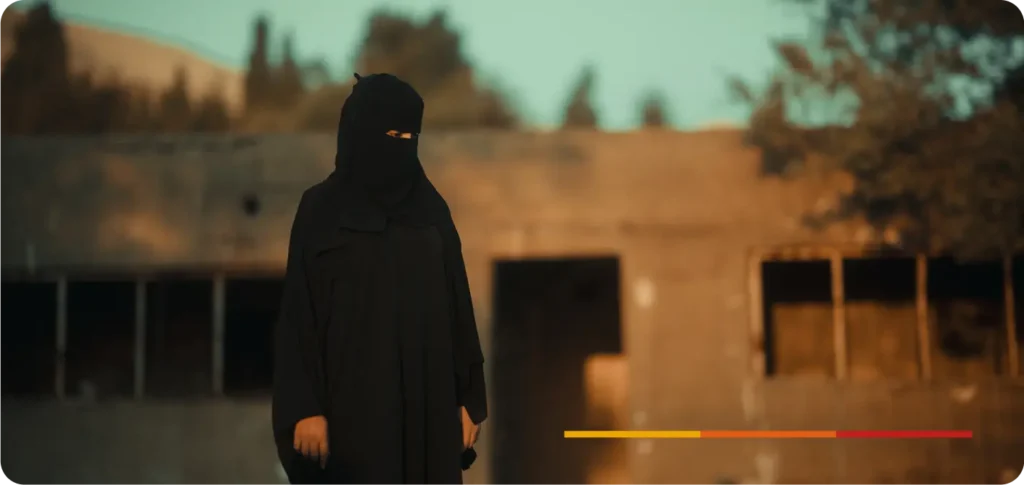 Mulher vestindo burca preta, com apenas os olhos visíveis, em pé diante de um prédio abandonado e danificado. Ao fundo, há vegetação e montanhas desfocadas, com iluminação de fim de tarde. No canto inferior direito, há uma barra colorida em tons de amarelo, laranja e vermelho.