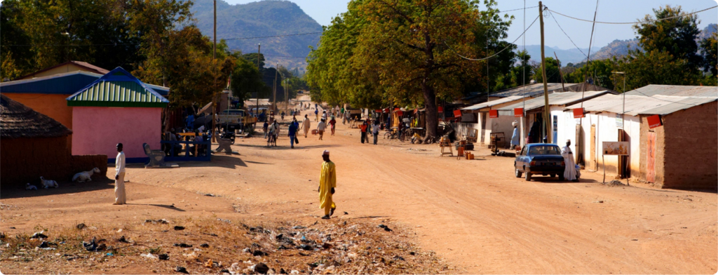 Rua de terra em uma vila africana, com pessoas caminhando, casas simples nas laterais, árvores ao fundo e montanhas ao longe. A cena retrata um dia ensolarado com céu claro e presença de comércio local.